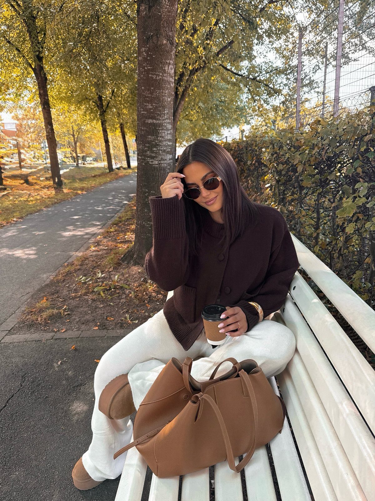 Woman in a dark brown sweater and white wide-leg pants sitting on a park bench.