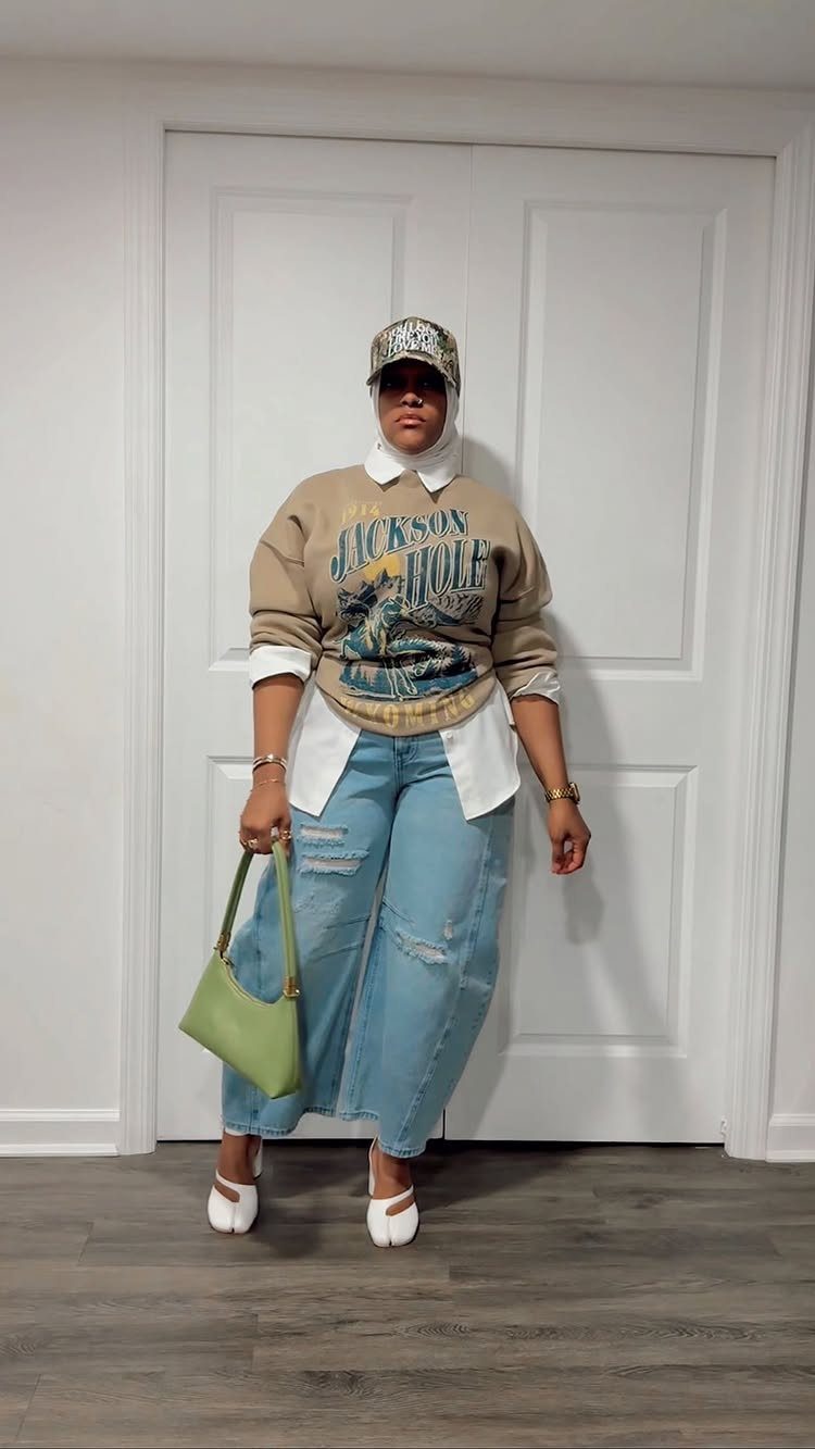 A person wearing a graphic sweater, white button-down, wide-leg jeans, and white Mary Jane shoes.