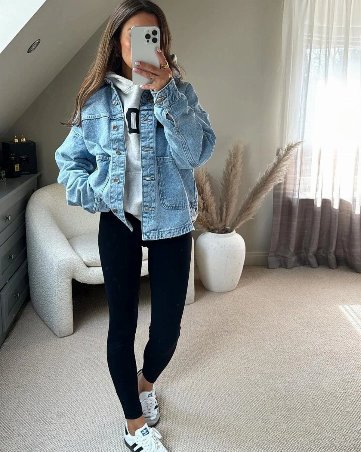 Woman in a denim jacket, grey hoodie, black leggings, and white sneakers taking a selfie.