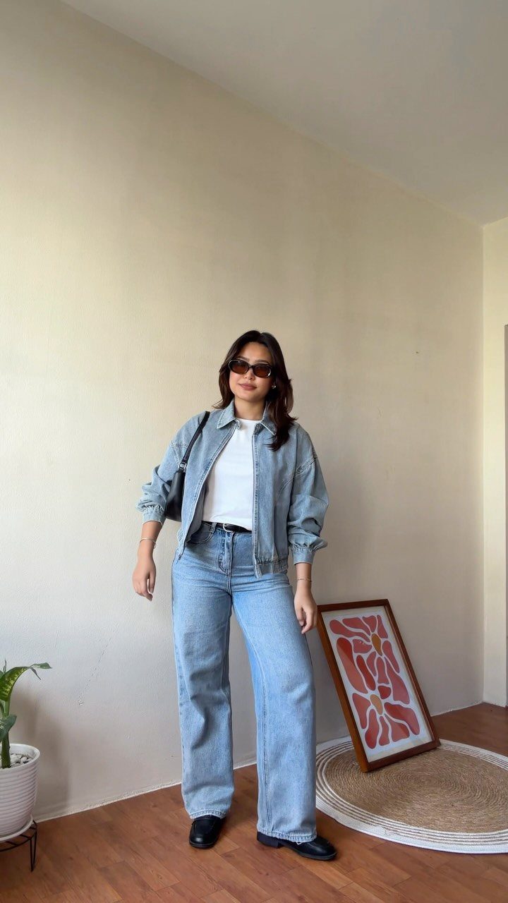 A woman wearing a denim jacket, white t-shirt, and wide-leg jeans standing indoors.