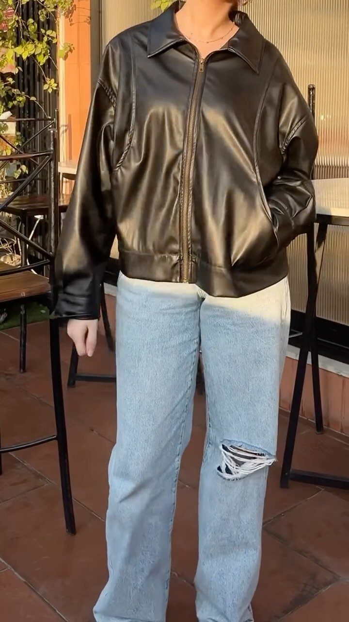 Person wearing a black leather jacket and light blue ripped wide-leg jeans standing outdoors.