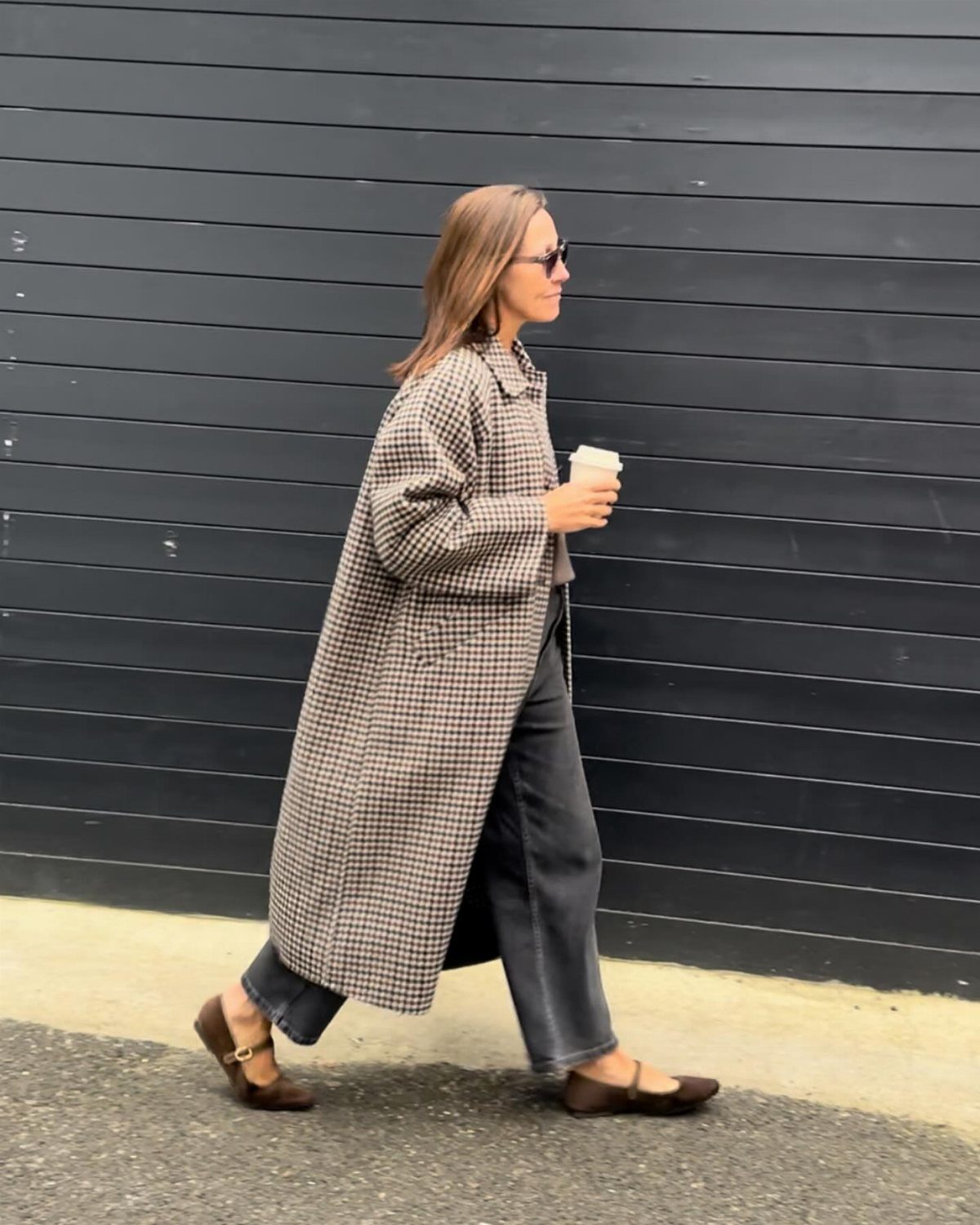 Woman walking in a long plaid coat, dark jeans, and brown ballet flats.