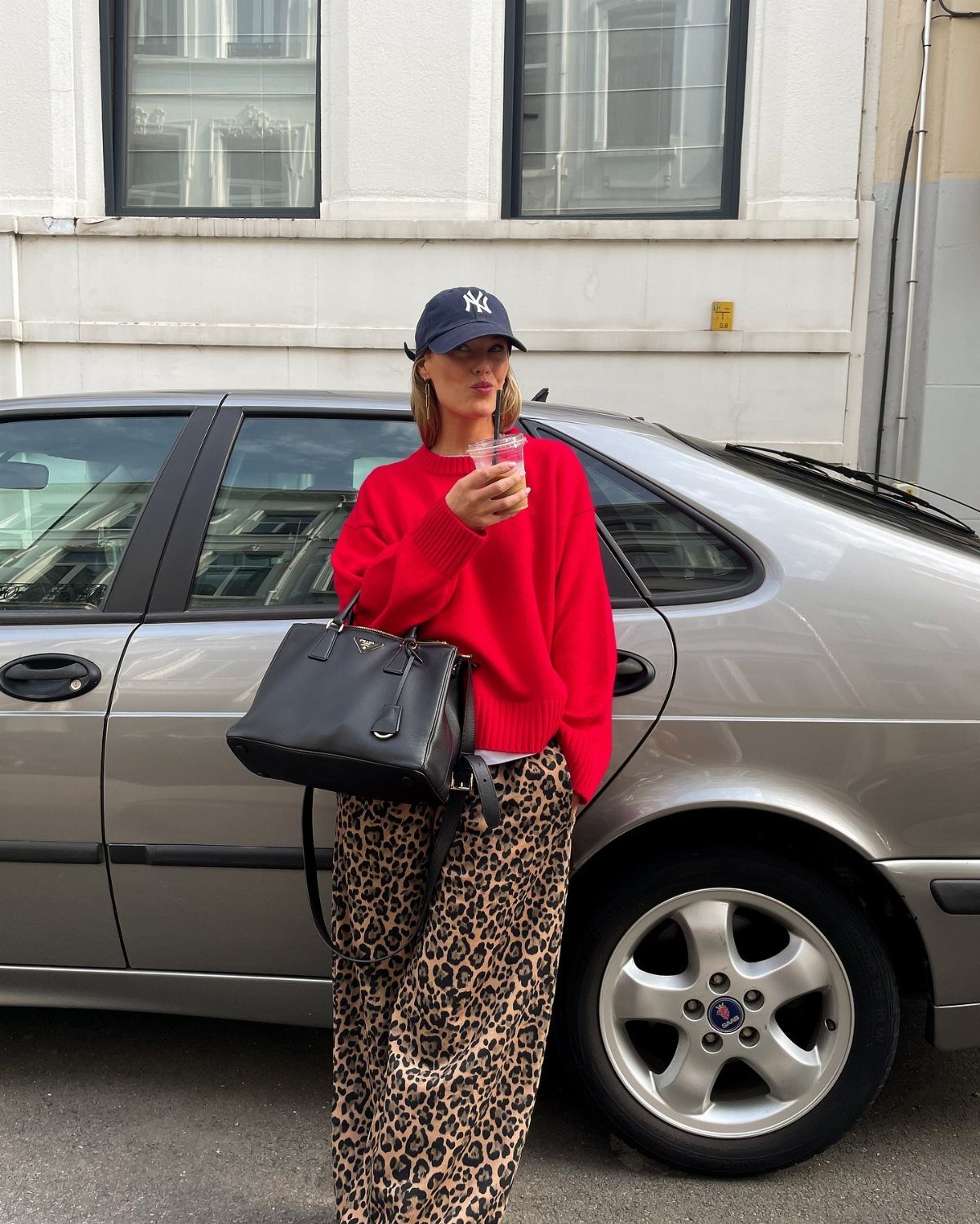Woman in a red sweater and leopard print pants standing by a silver car.