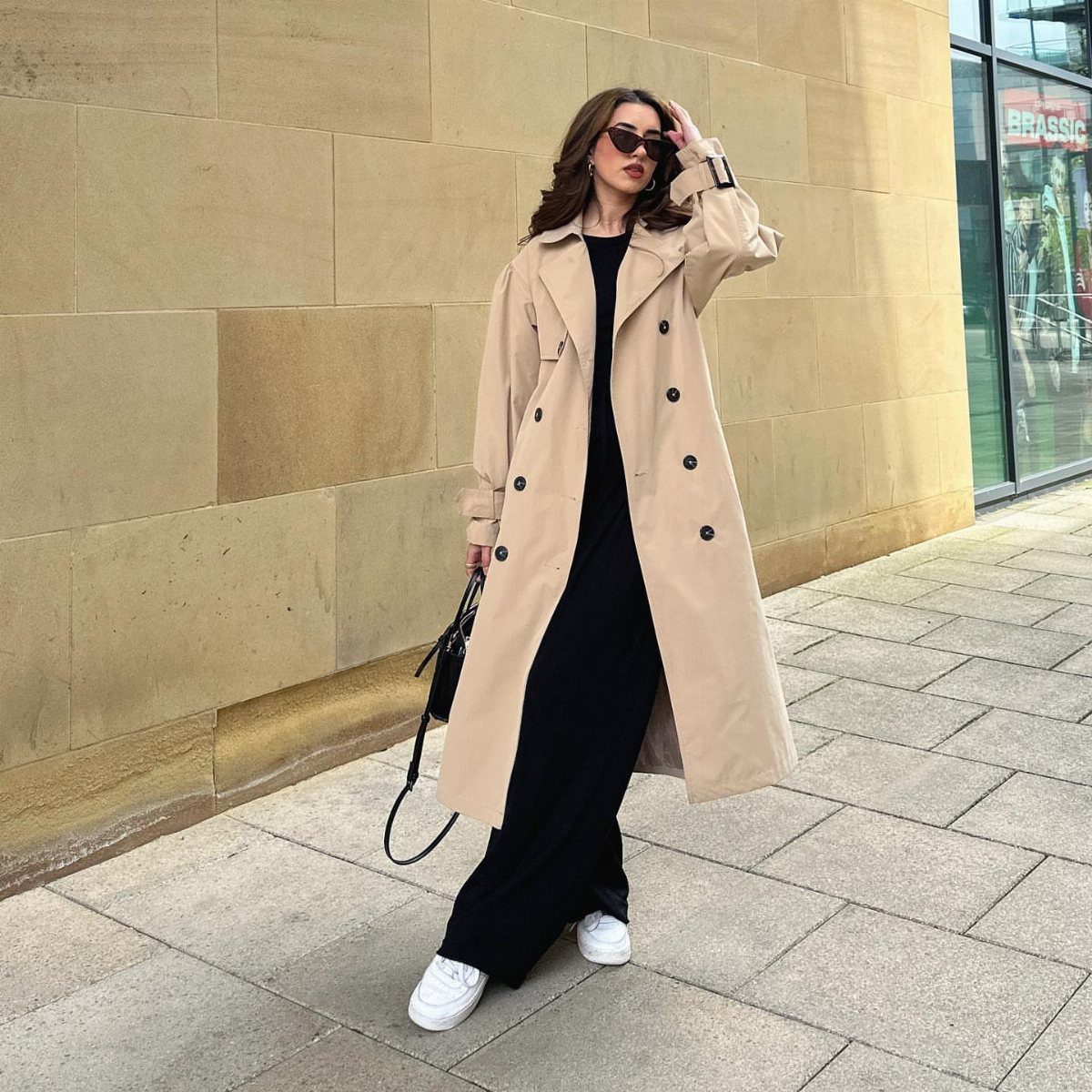 Woman in a long beige trench coat and black dress standing on a city sidewalk.