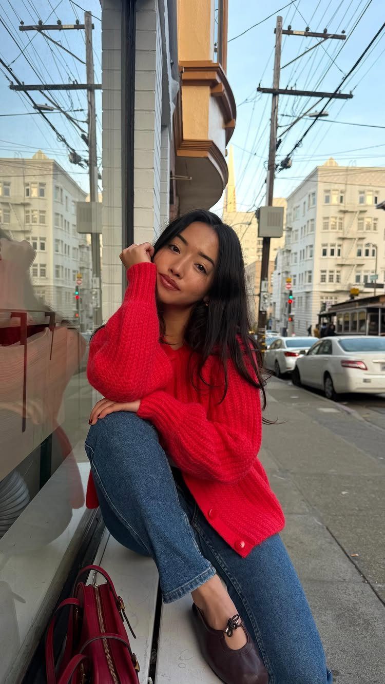 A woman wearing a bright red chunky knit sweater and blue jeans sitting on a city ledge.