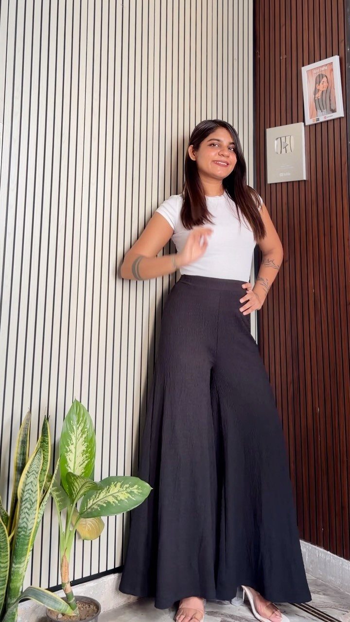 Woman wearing white sleeveless top and black high-waisted wide-leg trousers standing by a wall.