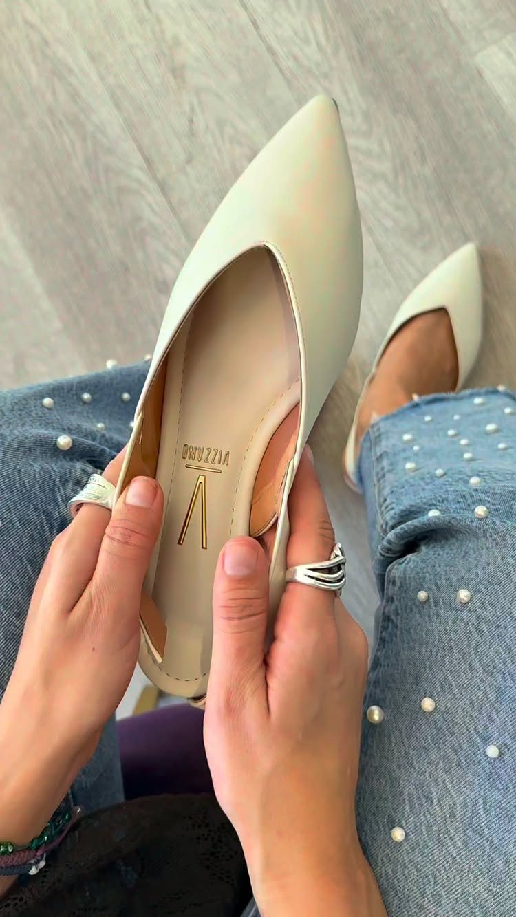 Cream pointed-toe flats held by hands over pearl-embellished blue denim jeans.
