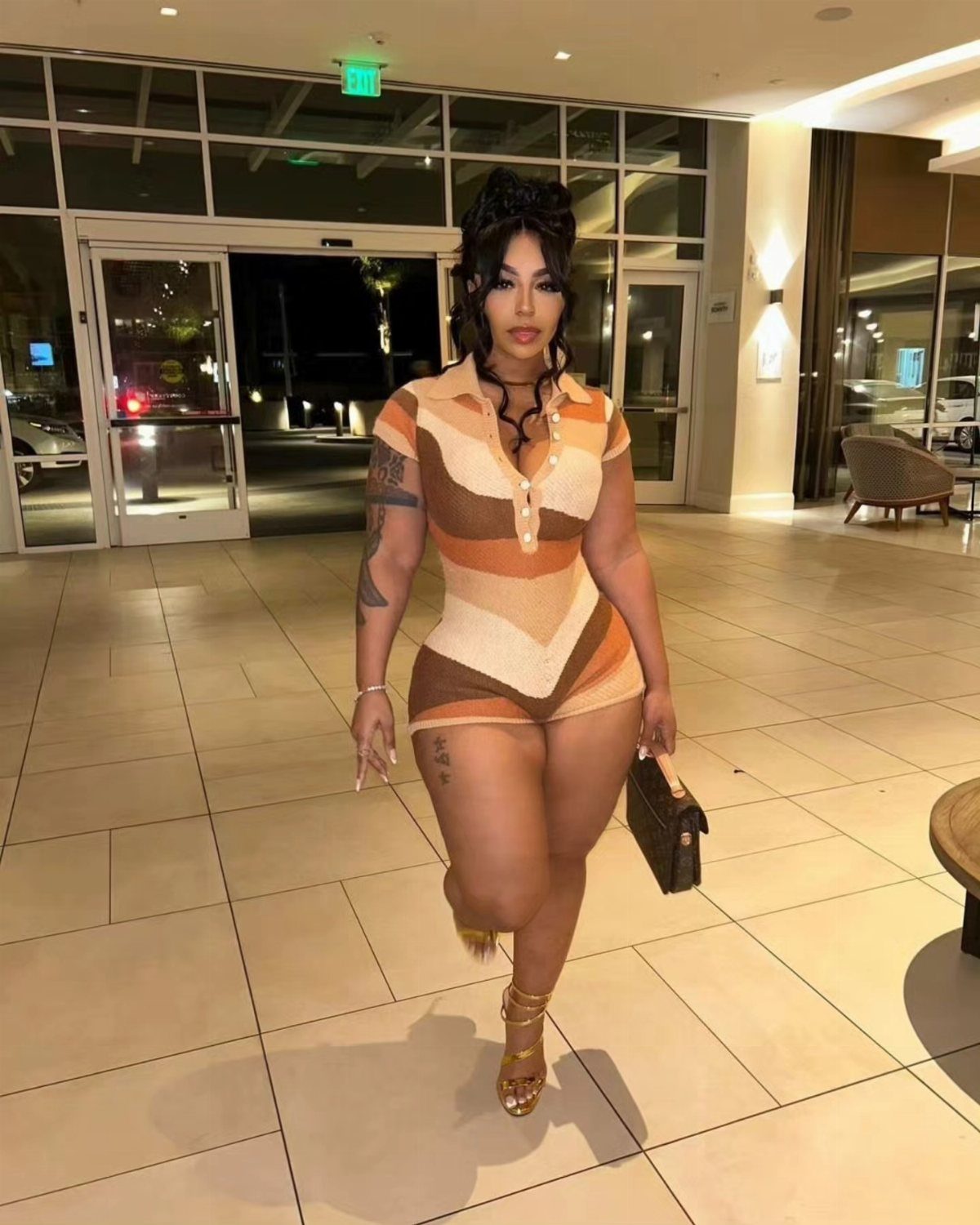 Woman in a patterned geometric romper walking through a modern building lobby wearing gold sandals.