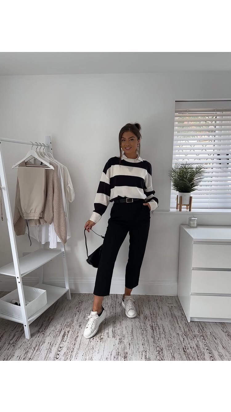 Woman in striped sweater and black trousers wearing white sneakers in a bright room.