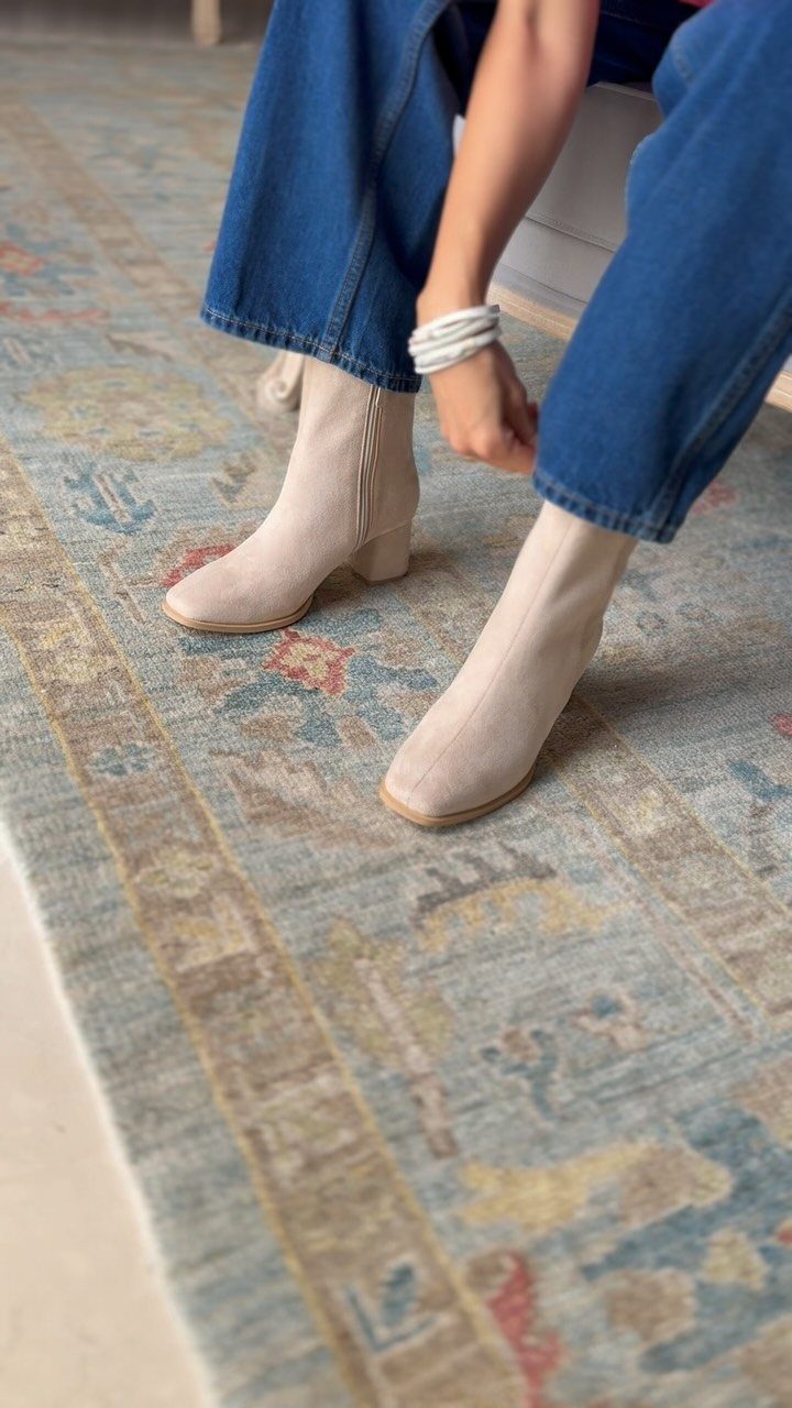 Beige suede ankle boots worn with blue denim jeans on a patterned rug.