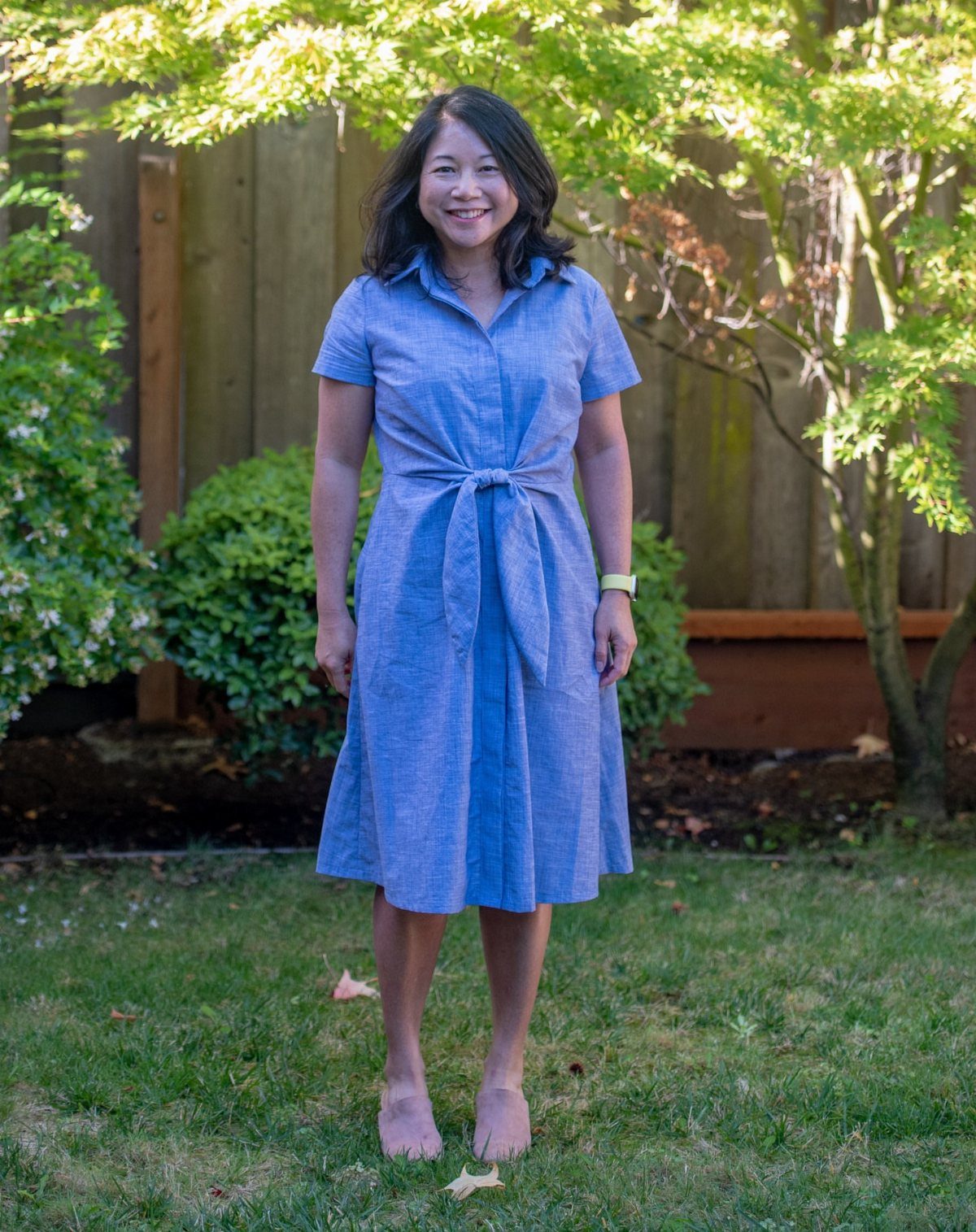 A smiling woman wearing a blue short-sleeved chambray shirtdress standing in a garden.