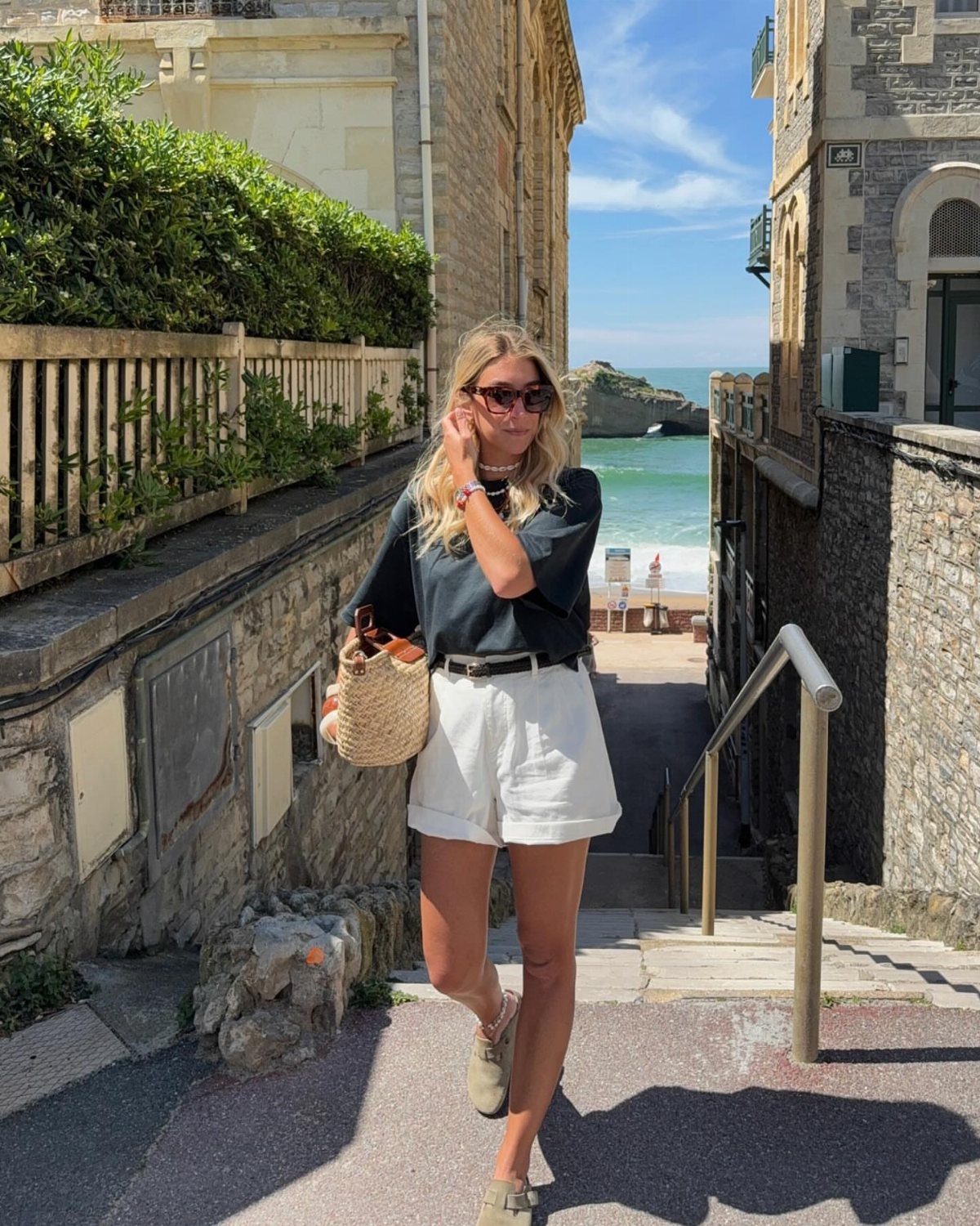 Woman wearing a dark top and white linen shorts standing on outdoor stairs by the sea.