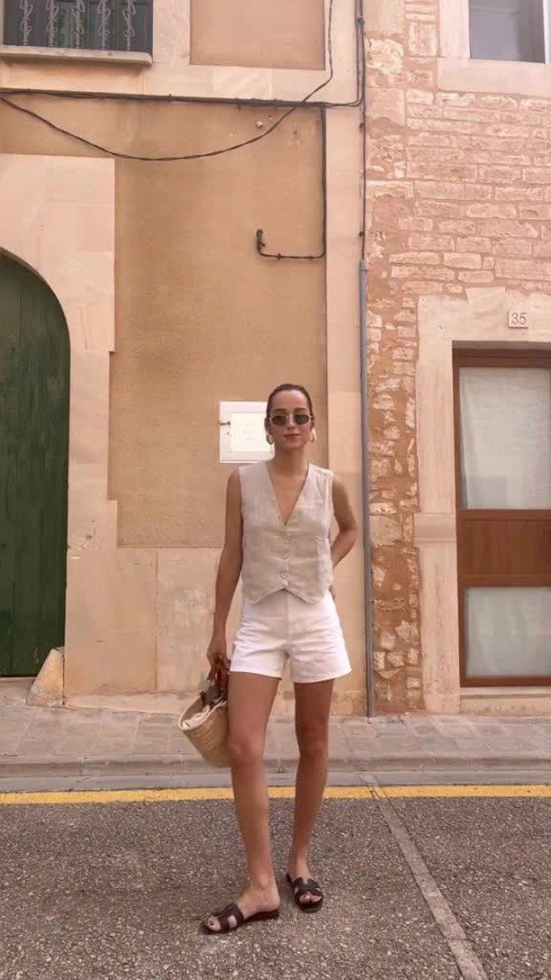 Woman wearing a beige vest and white shorts standing on a street by stone buildings.