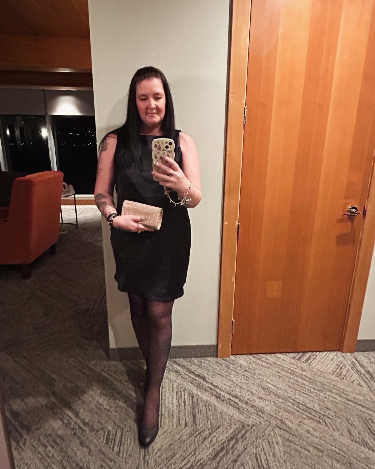 Woman in a short black dress, black tights, and heels holding a beige clutch.
