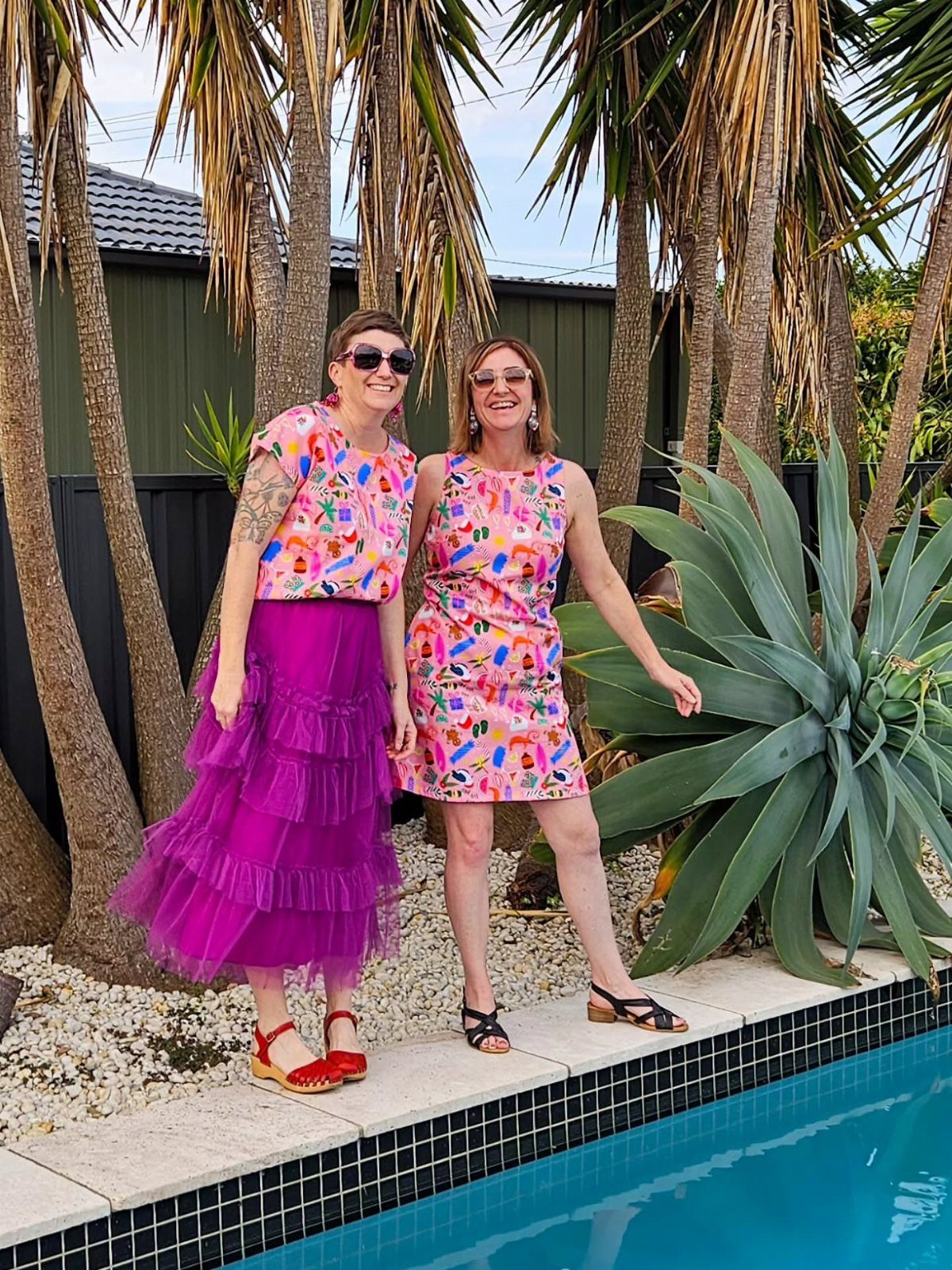 Two women posing in colorful festive summer outfits by a swimming pool with palm trees.