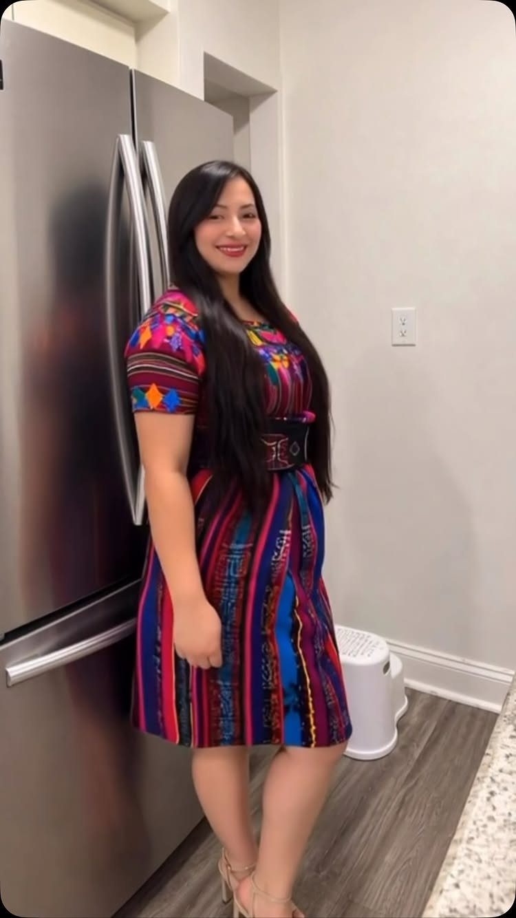 A smiling woman wearing a vibrant, colorful patterned short-sleeved dress standing in a kitchen.