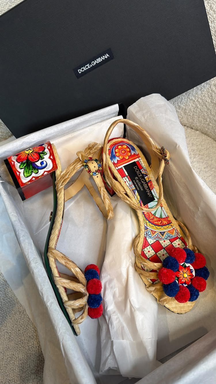 Woven straw sandals with colorful patterns and red and blue pom-poms inside a designer box.