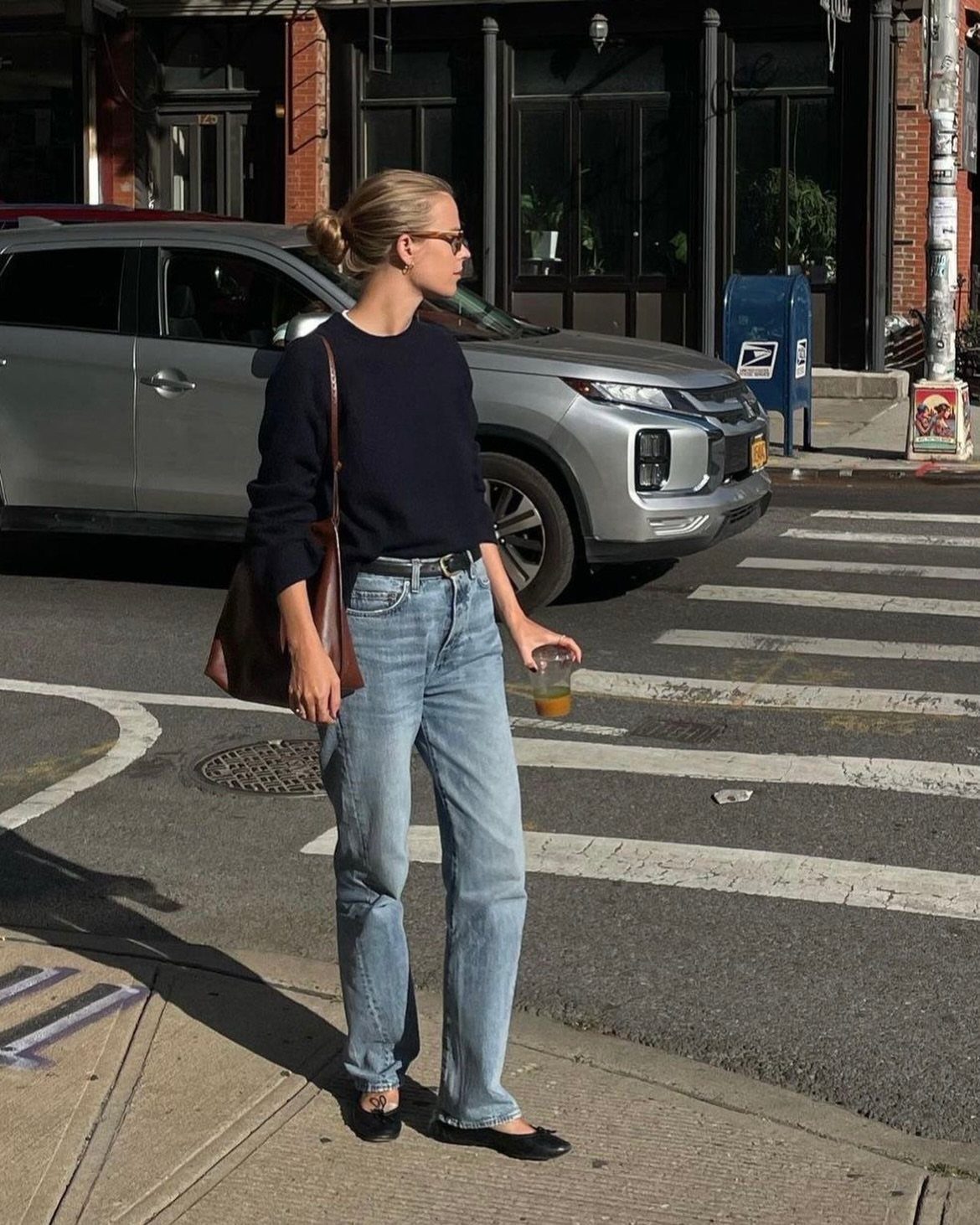 A woman in a navy sweater and light blue jeans standing on a city street corner.