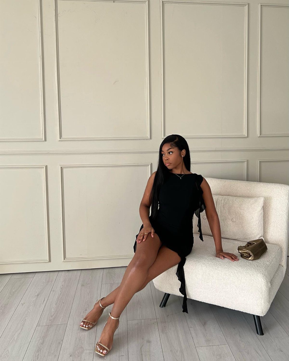A woman in a black mini dress and gold heels sitting on a white armchair.
