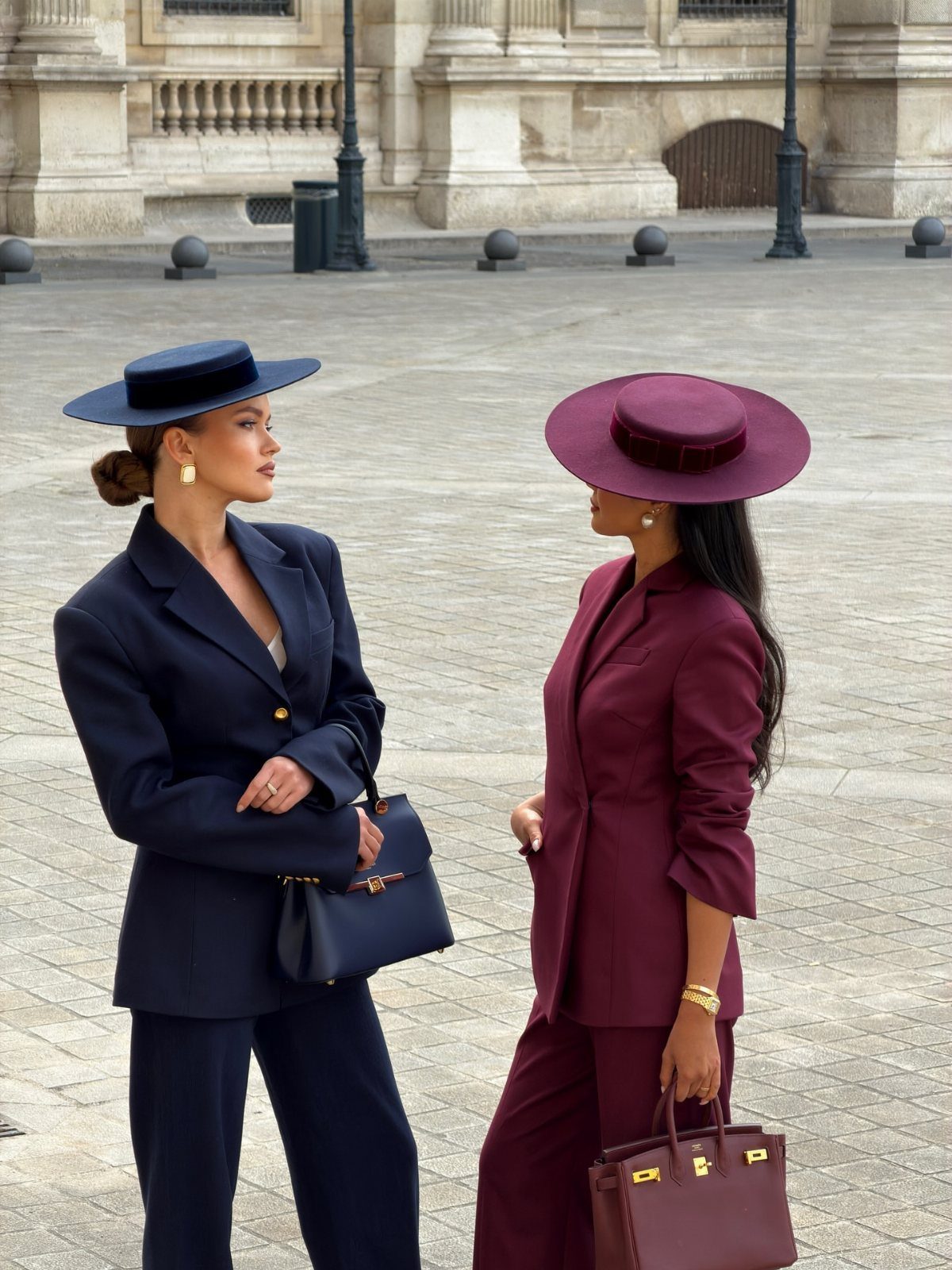 Two women in elegant monochromatic tailored suits and wide-brimmed hats standing in a city plaza.