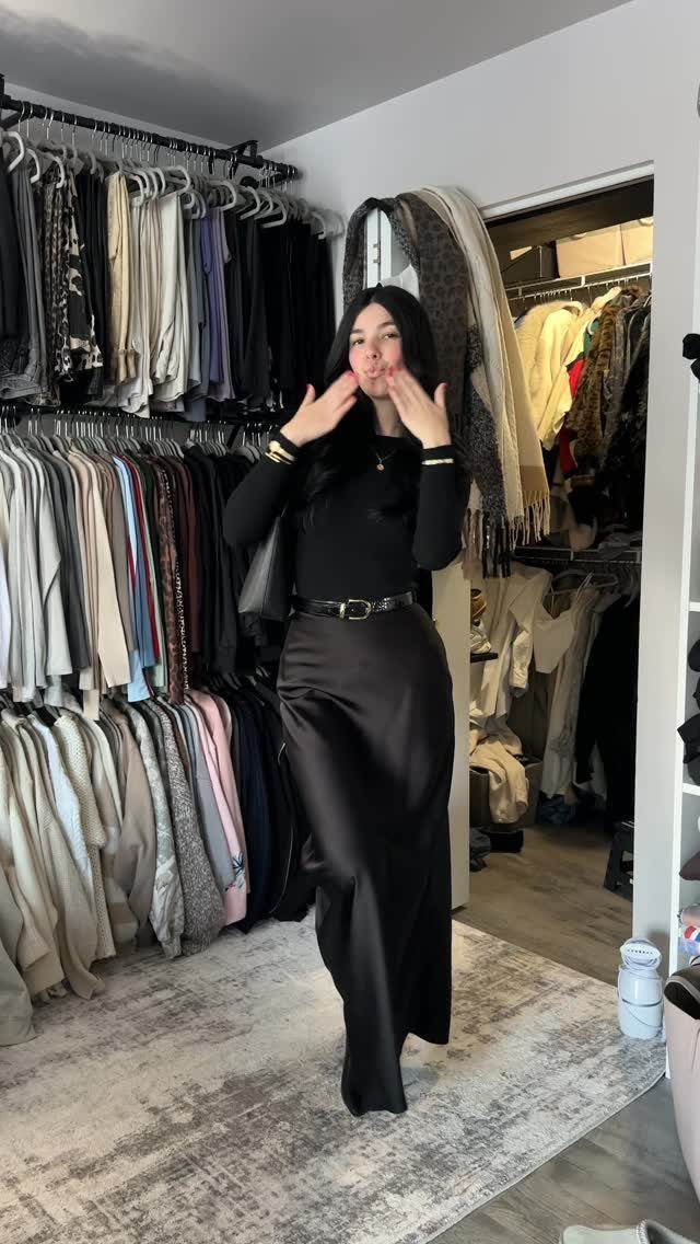 Woman in a black long-sleeved top and black satin midi skirt standing in a closet.