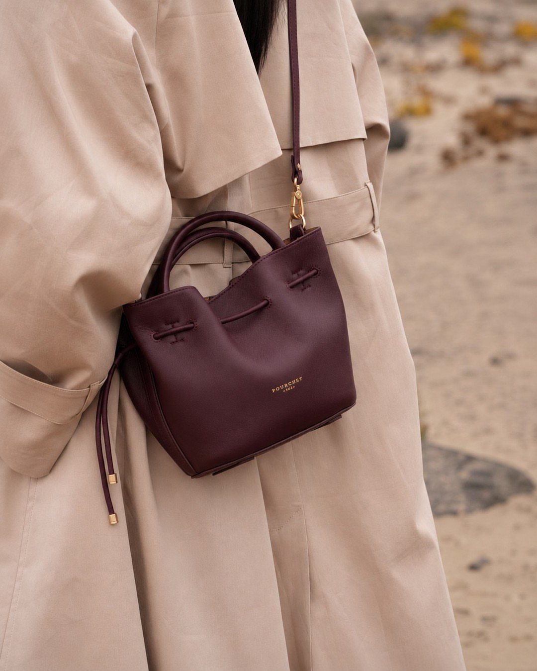 A burgundy structured leather handbag worn over a beige trench coat on a sandy beach.