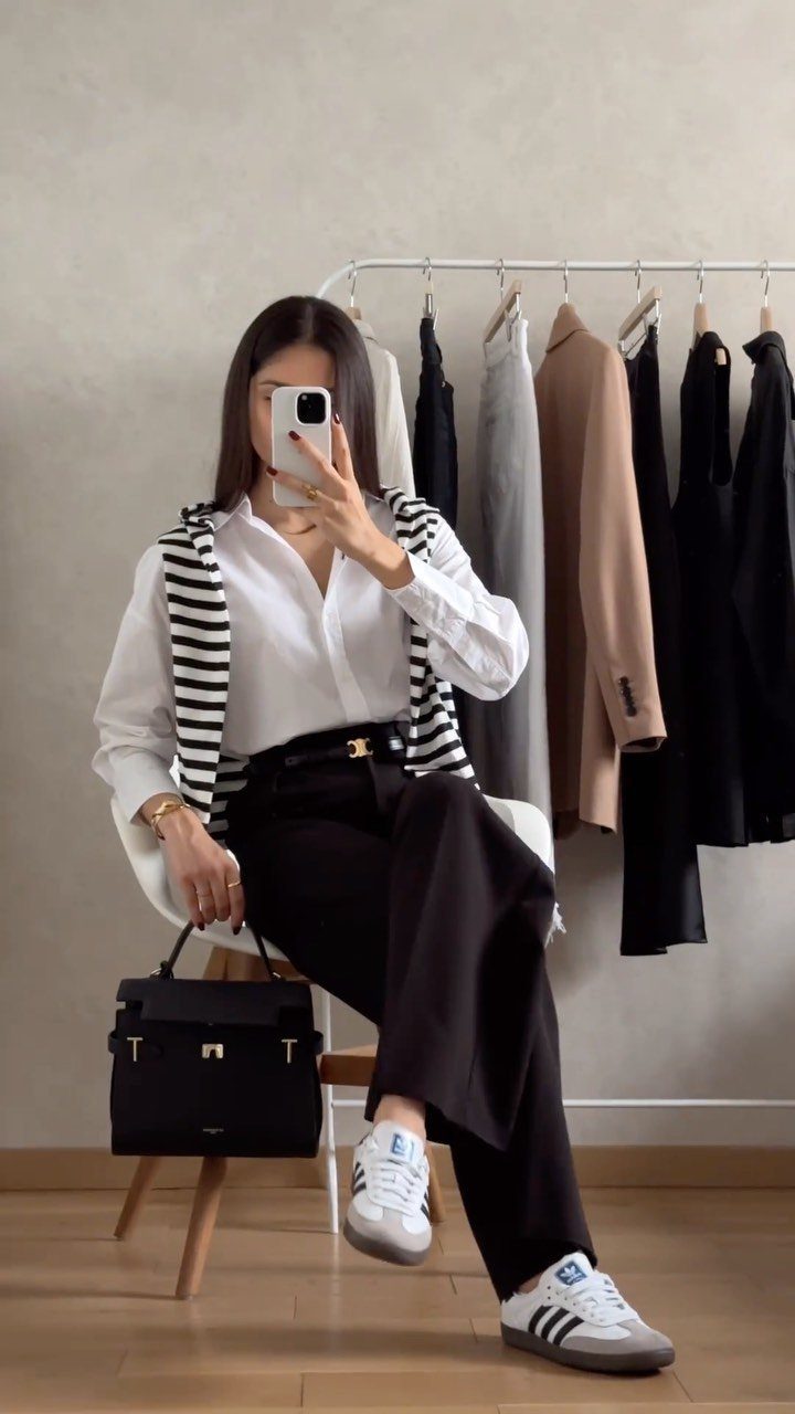 A woman posing in a white shirt, striped sweater, and black trousers while taking a selfie.