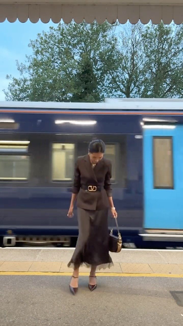 A woman in a dark brown belted blazer and midi skirt standing on a train platform.
