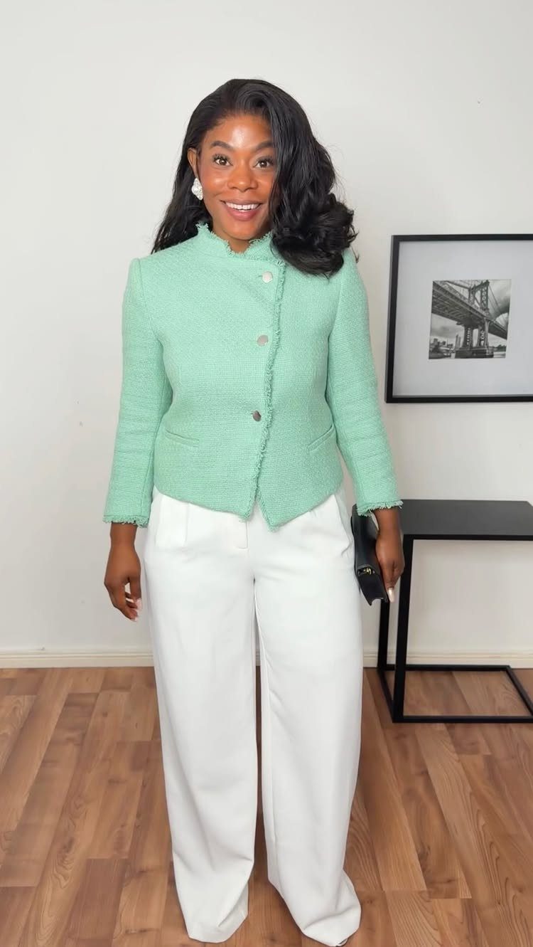 A smiling woman wearing a mint green jacket and white wide-leg trousers standing indoors.