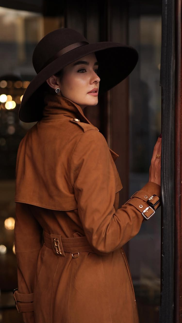 A woman in a wide-brimmed hat and brown belted trench coat looking over her shoulder.