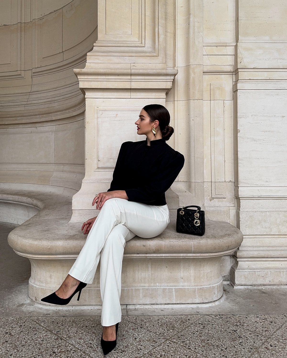 Woman in a black top and white trousers sitting on a stone ledge outdoors.