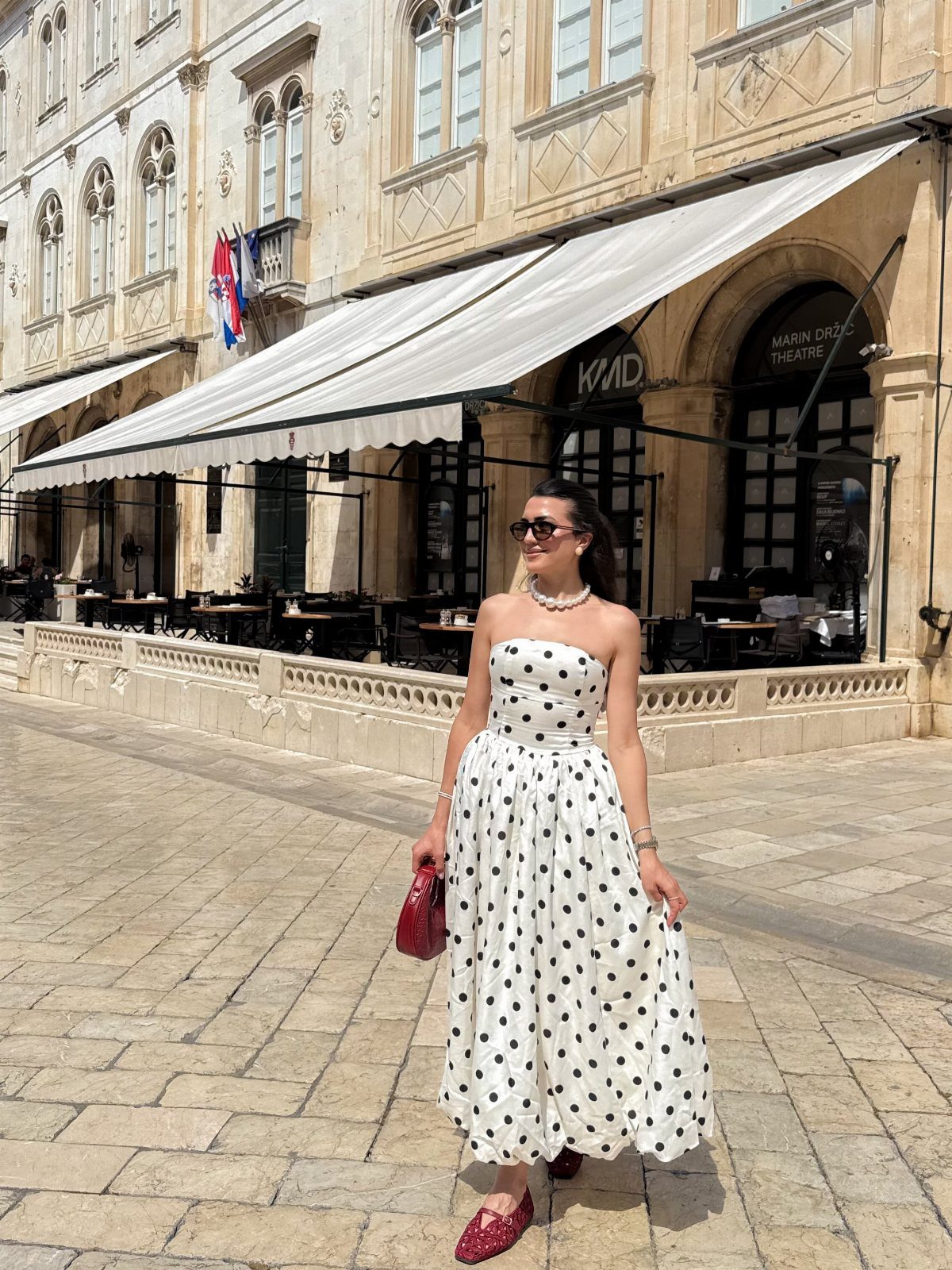 Woman in a white polka dot strapless dress standing on a sunny cobblestone street.