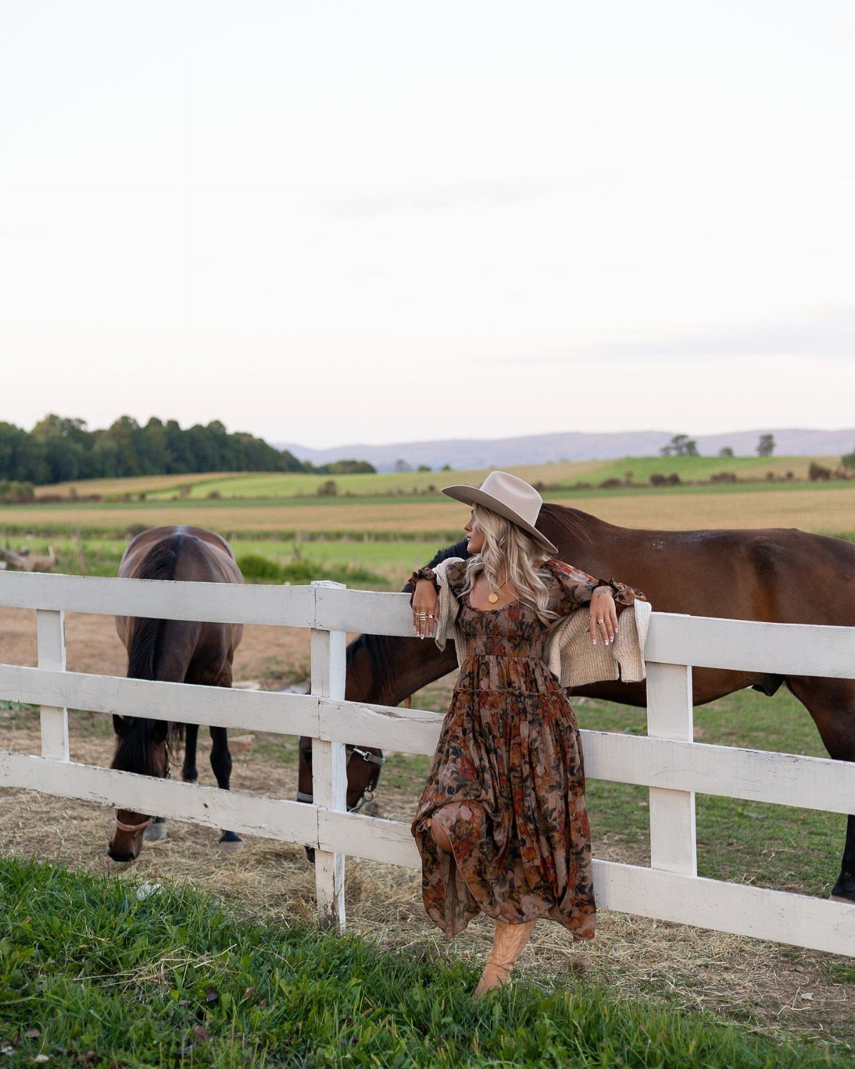 Woman in a brown floral print dress leaning against a white fence in a pasture.