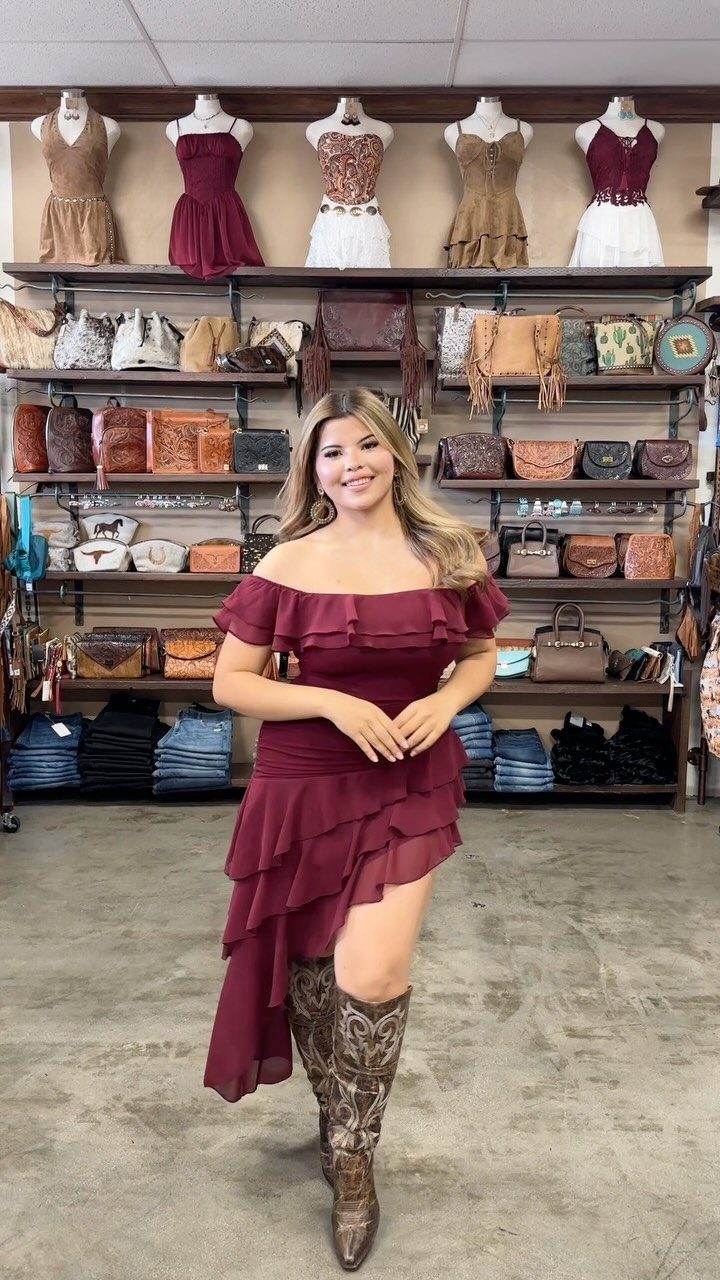 A woman wearing a maroon off-the-shoulder ruffled high-low dress and cowboy boots.