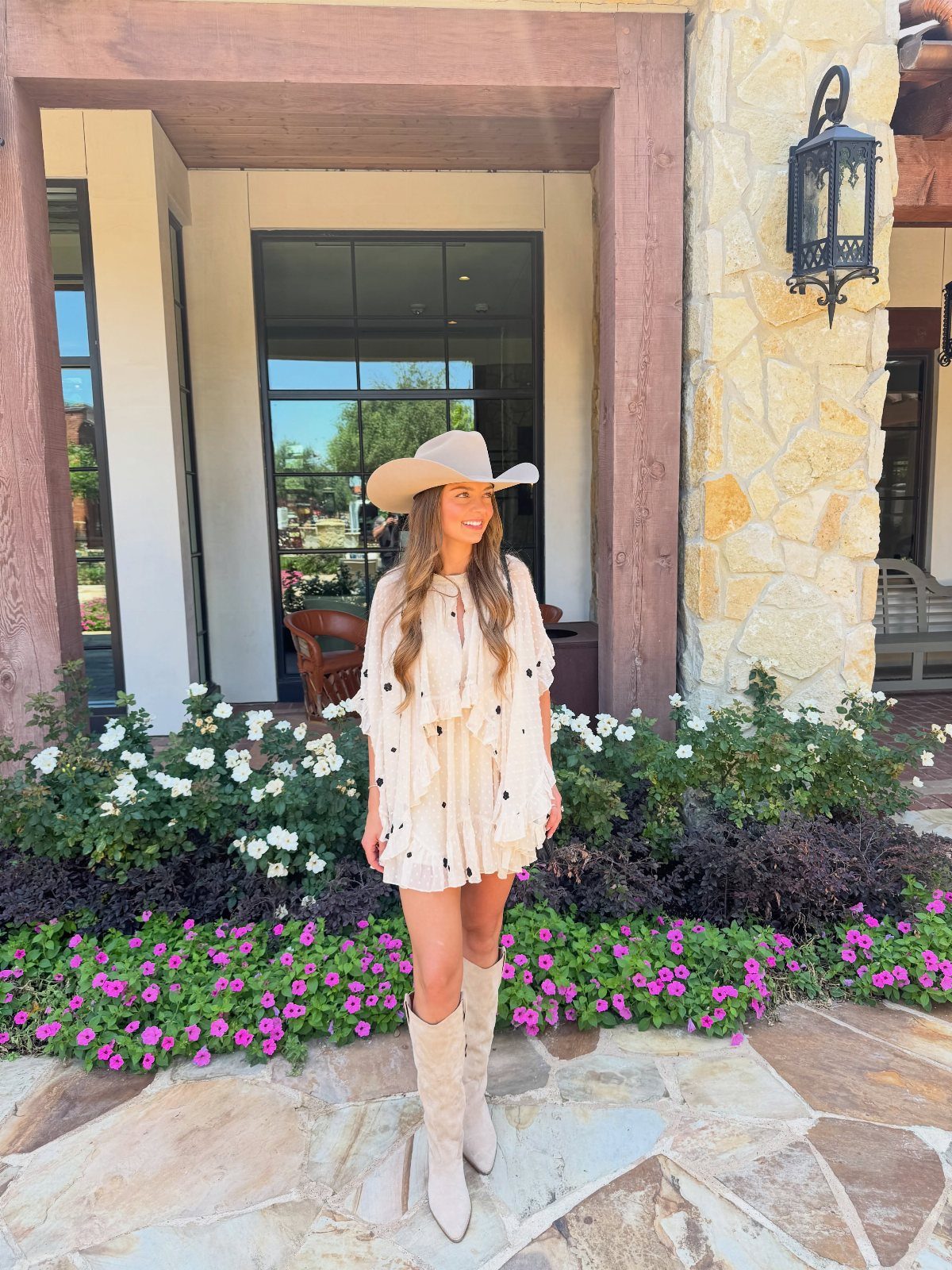 Woman in a cream ruffled polka dot dress, white cowboy hat, and light boots standing outdoors.