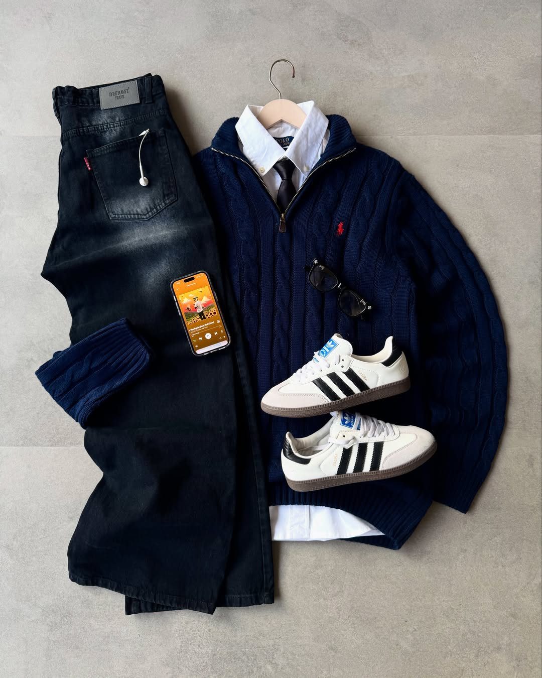 Navy cable-knit sweater, white collared shirt, dark jeans, and white sneakers arranged in a flat lay.