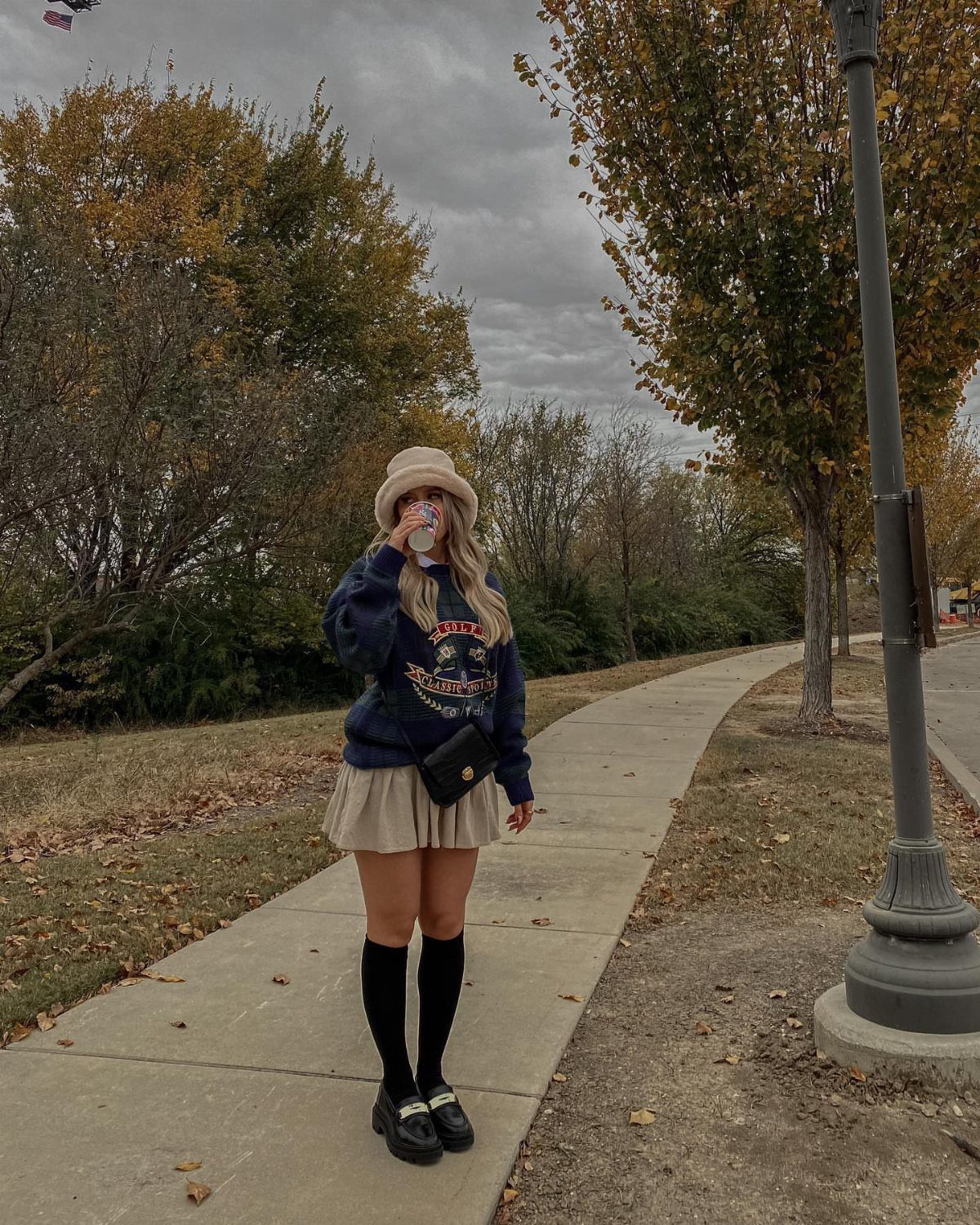 A person wearing a navy sweatshirt, beige pleated skirt, black knee-high socks, and black loafers standing outdoors.