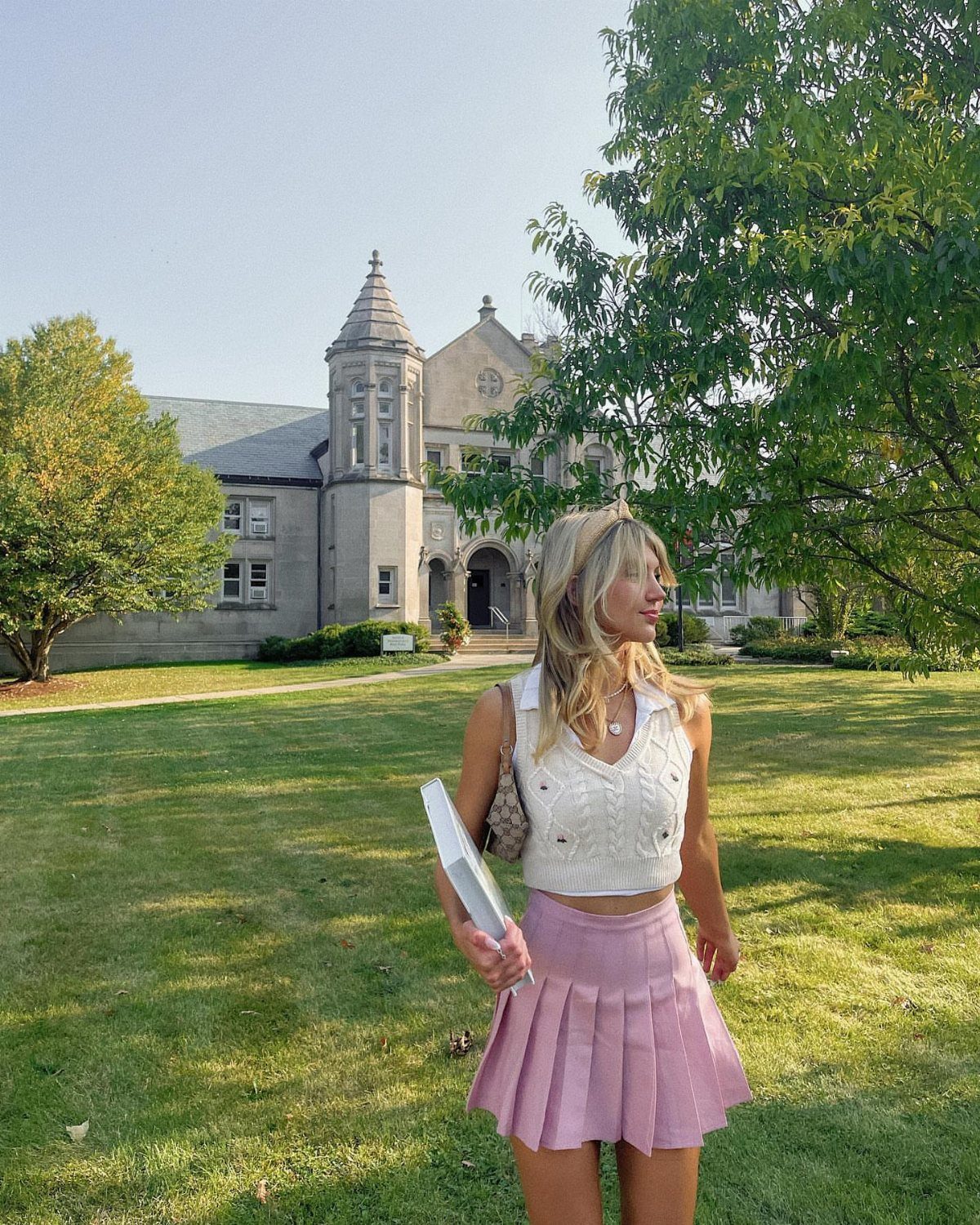 A woman wearing a white cable-knit sweater vest and a pink pleated skirt standing on a lawn.