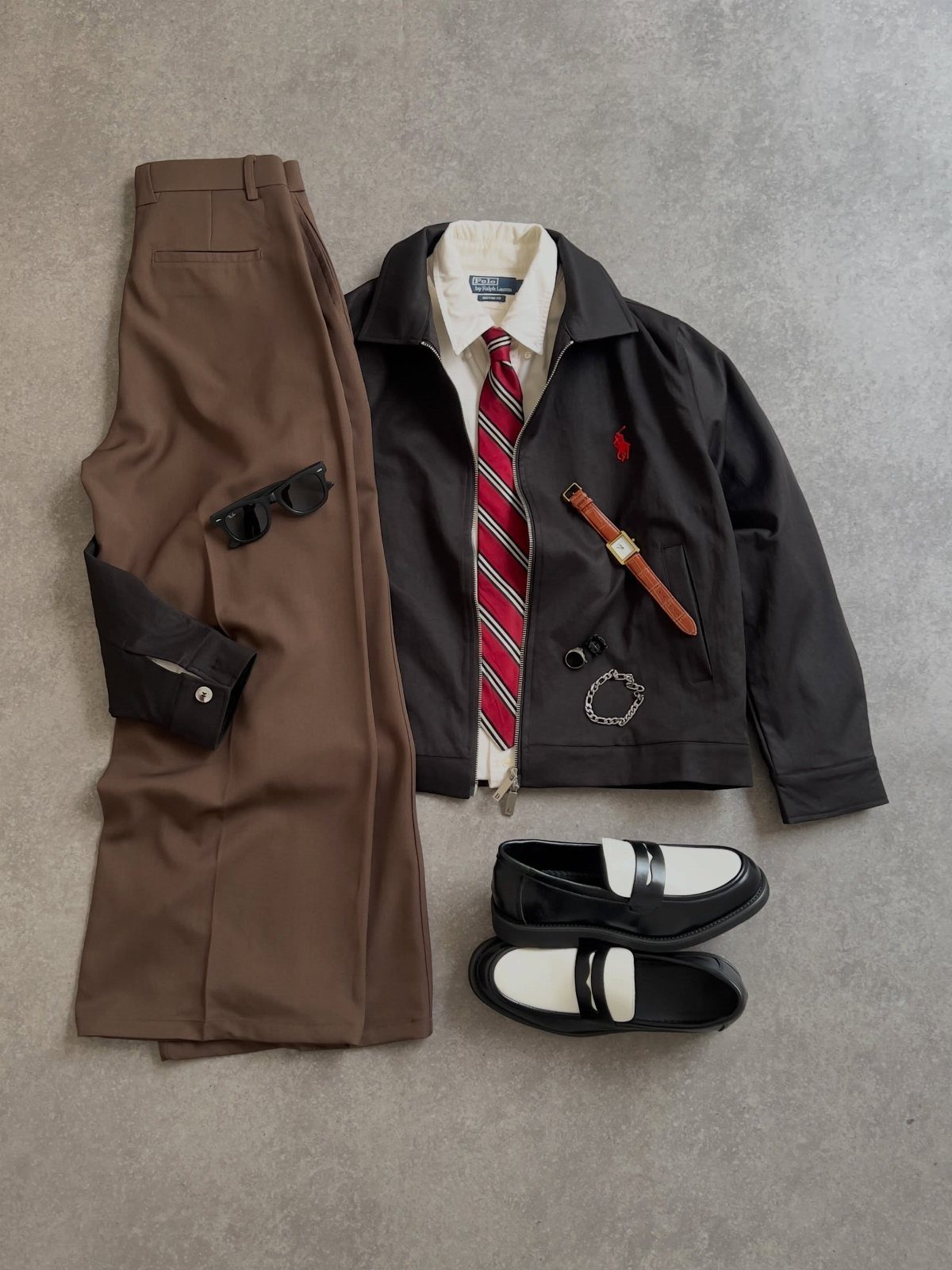 Flat lay of brown trousers, white shirt, red striped tie, black jacket, and loafers on grey background.