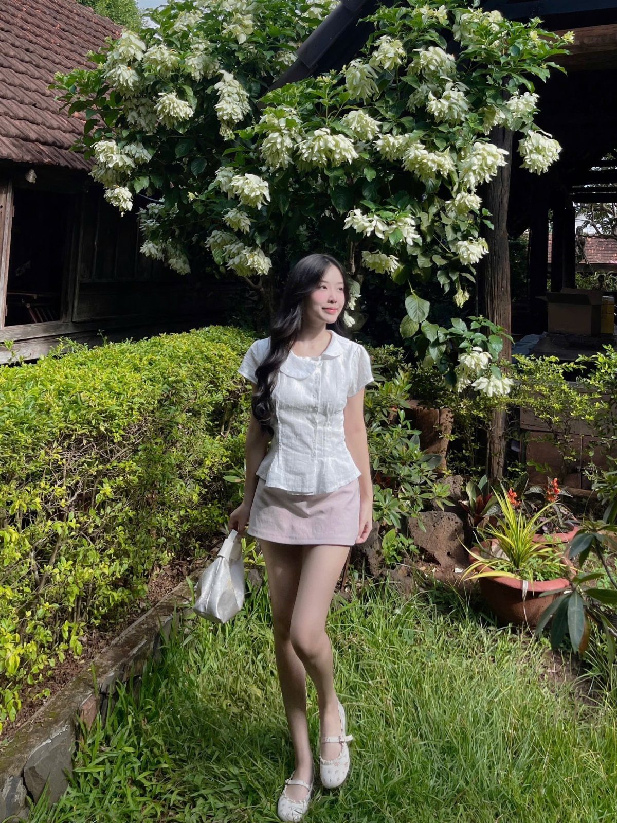 A young woman in a white top and pink mini skirt standing in a garden.