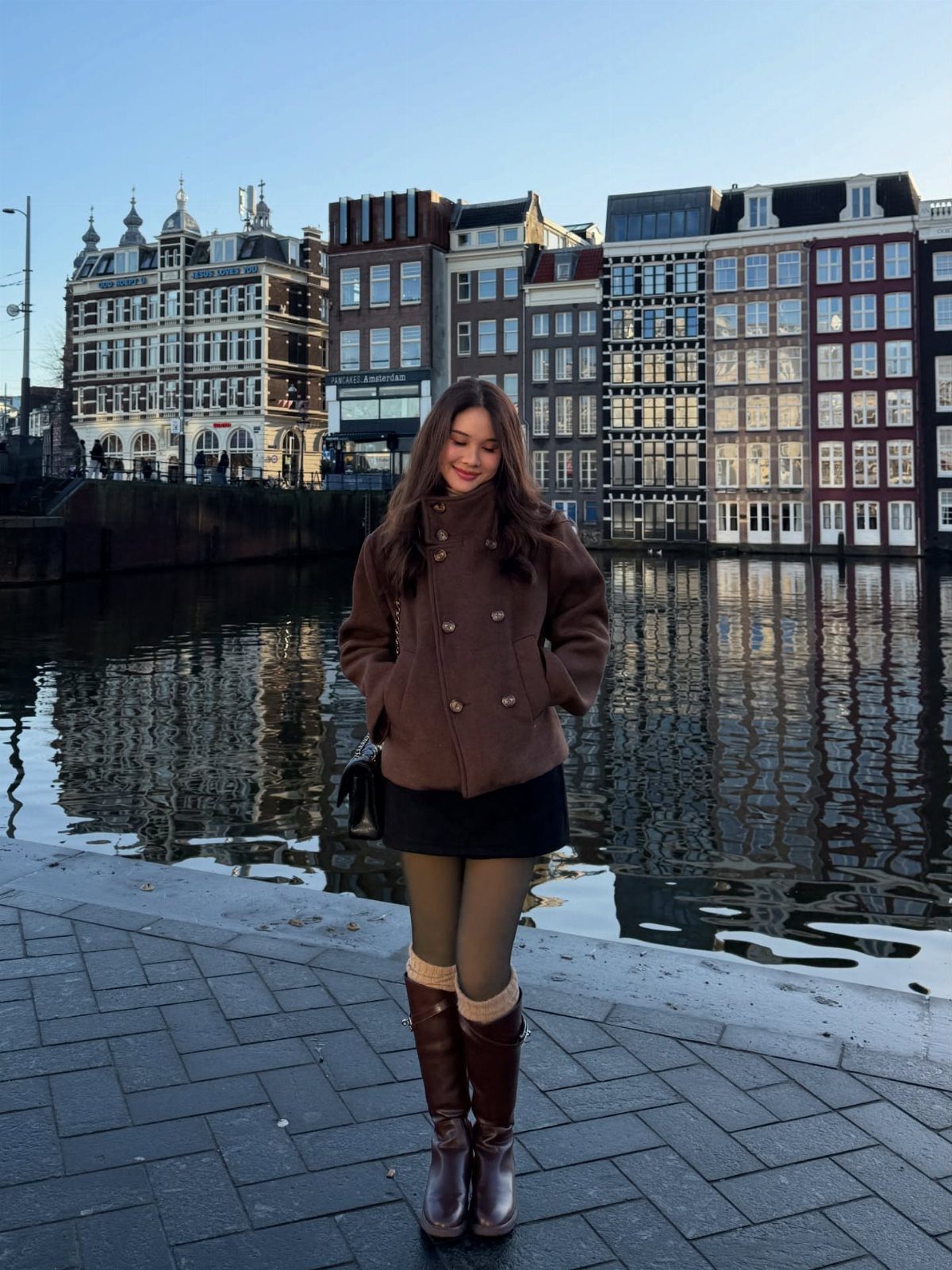 A woman wearing a brown jacket, black skirt, green tights, and tall boots by a canal.