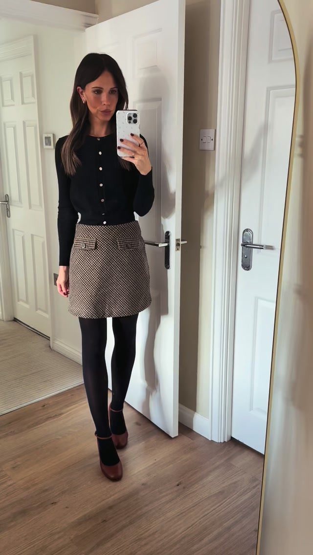 Woman in a black top, patterned mini skirt, black tights, and flats posing for a mirror selfie.