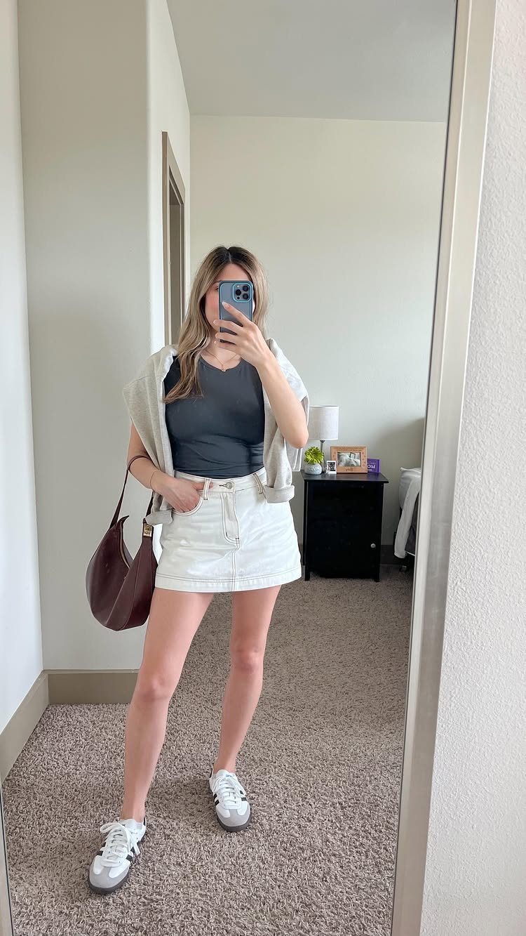 Woman in a white denim mini skirt, grey tank top, and white sneakers posing for a mirror selfie.