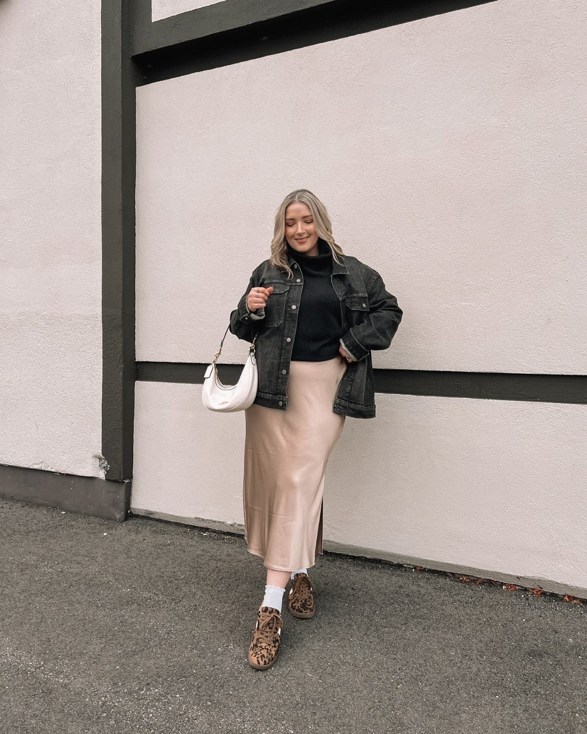 A woman wearing a black turtleneck, denim jacket, and beige midi skirt standing outdoors.