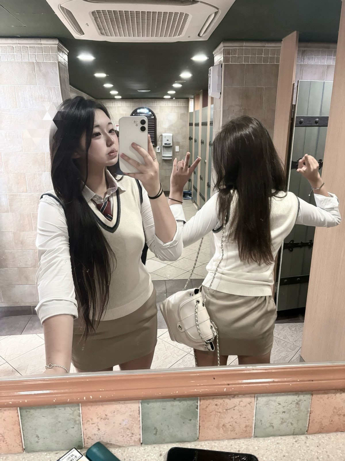 Woman in a cream sweater vest and tan mini skirt taking a mirror selfie.