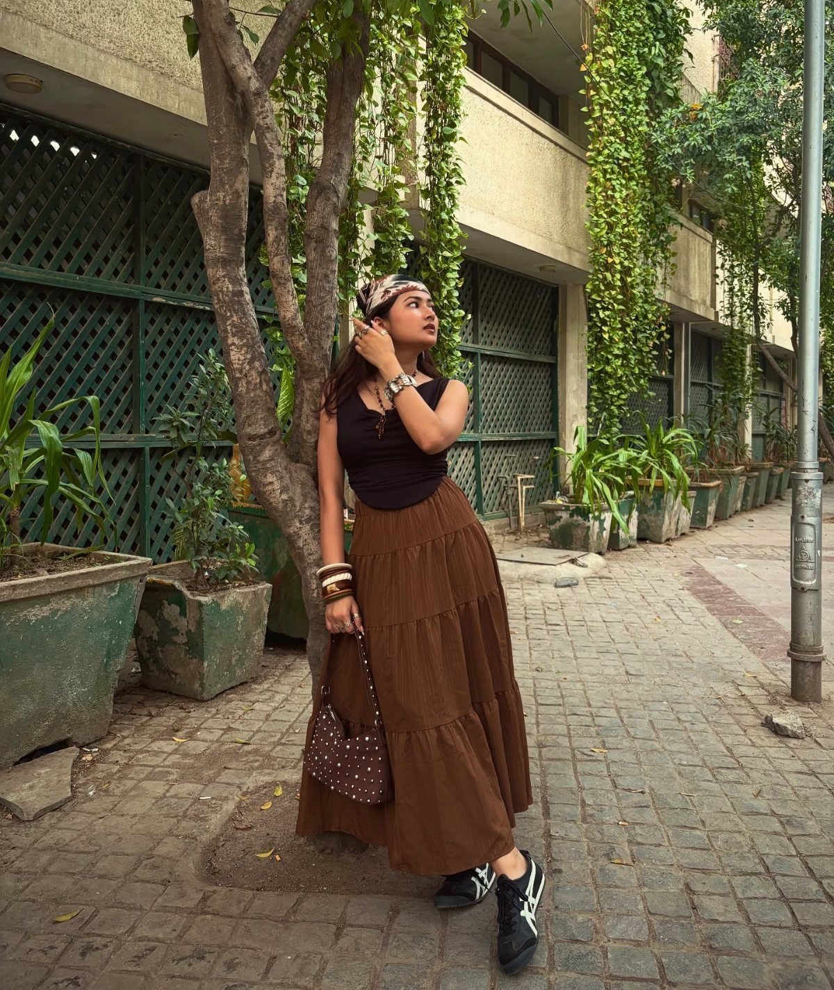 A woman wearing a long brown tiered skirt and black top standing near green plants.