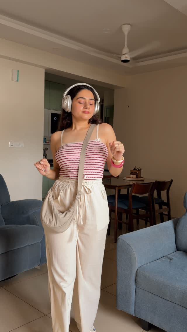 A woman in a striped tank top and cream flowy pants wearing headphones indoors.