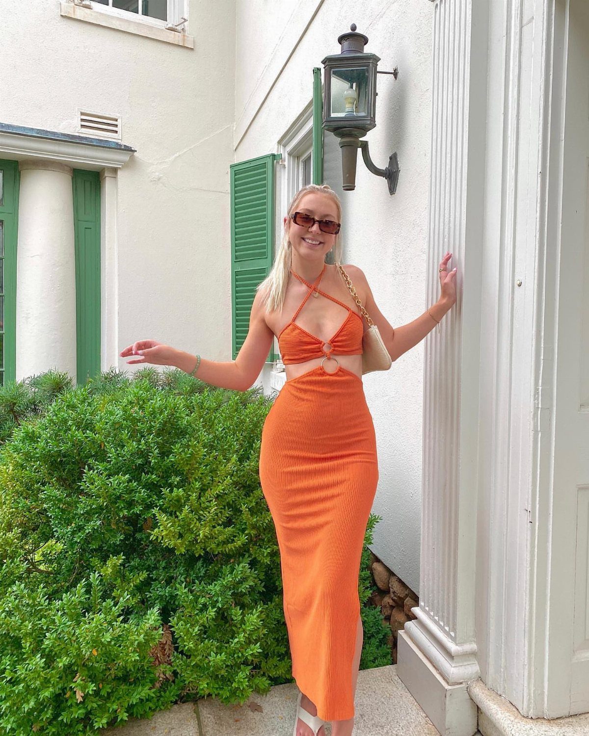 Blonde woman in an orange ribbed midi dress posing next to a white building and green bushes.
