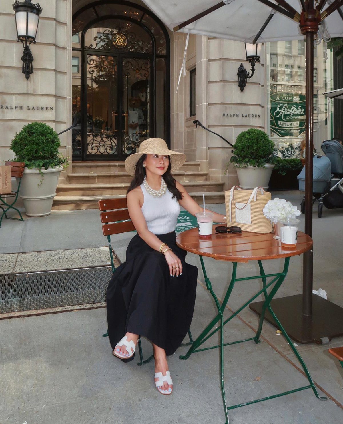 A woman in a white top and black skirt sits at a cafe wearing a straw hat.