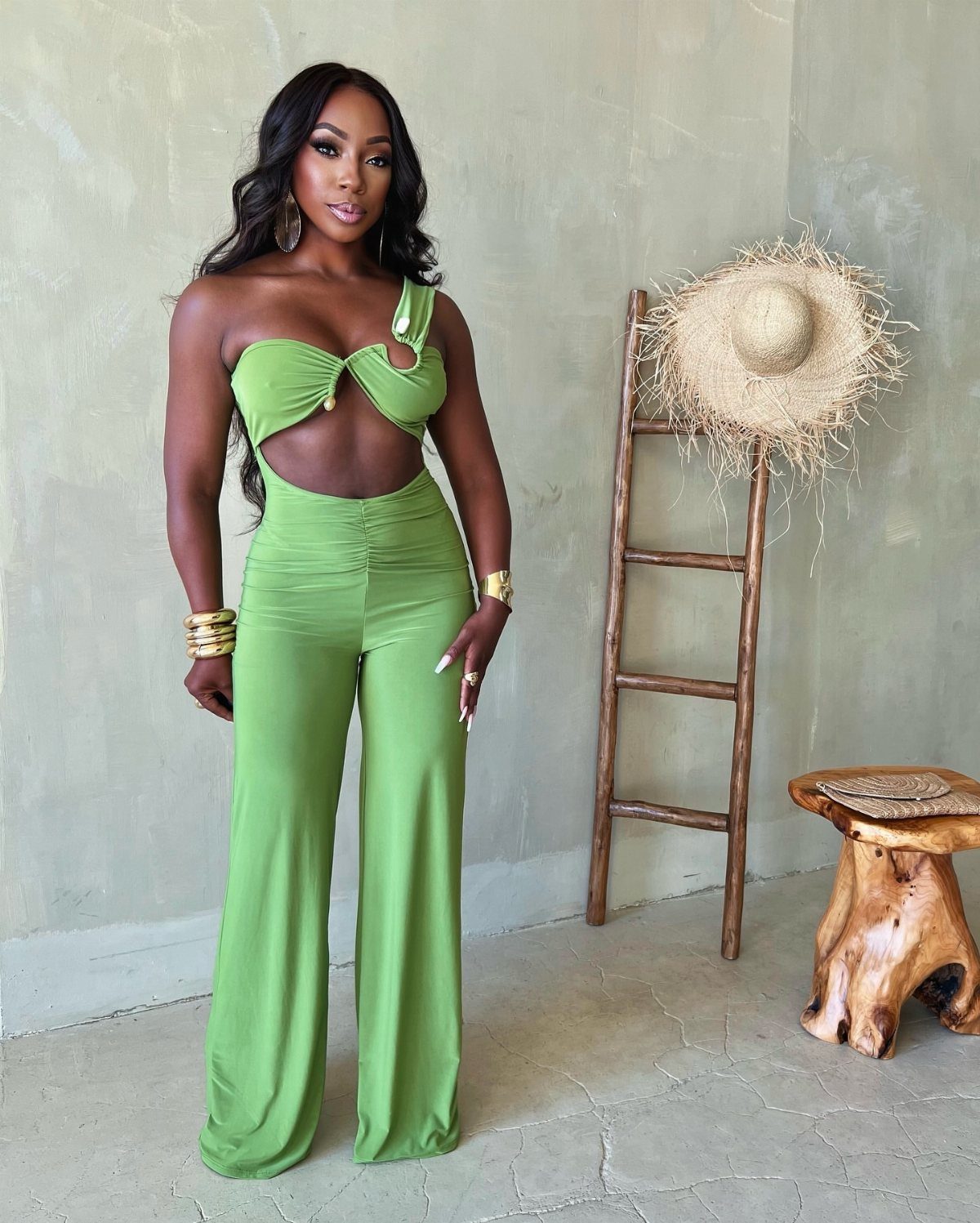 A woman posing in a lime green one-shoulder cutout jumpsuit against a neutral wall.